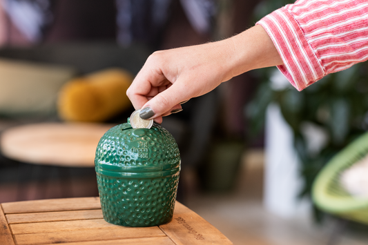 De spaarpot staat op een acacia zijplankje van een Big GreenEgg. Een dameshand stopt een muntje in de sleuf van de spaarpot.