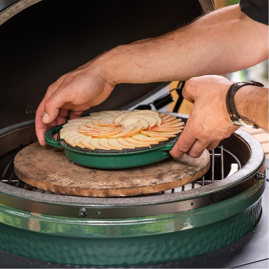 Dezelfde steen gelegen op een RVS rooster in een Big Green Egg. Op de baking stone staat de groene deksel van een Big Green Egg Dutch Oven met daarin appelplakjes in waaiervorm neergelegd.