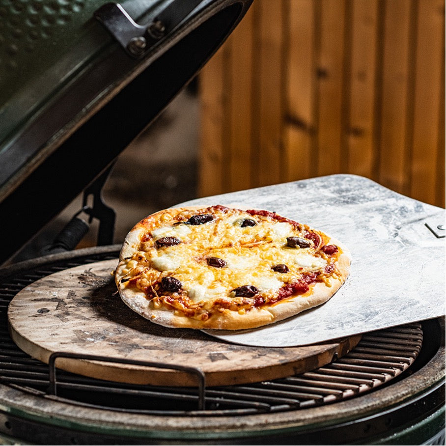 Dezelfde steen gelegen op een RVS rooster in een Big Green Egg. Op de steen ligt een pizzaschep met daarop een heerlijk uitziende, afgebakken pizza.