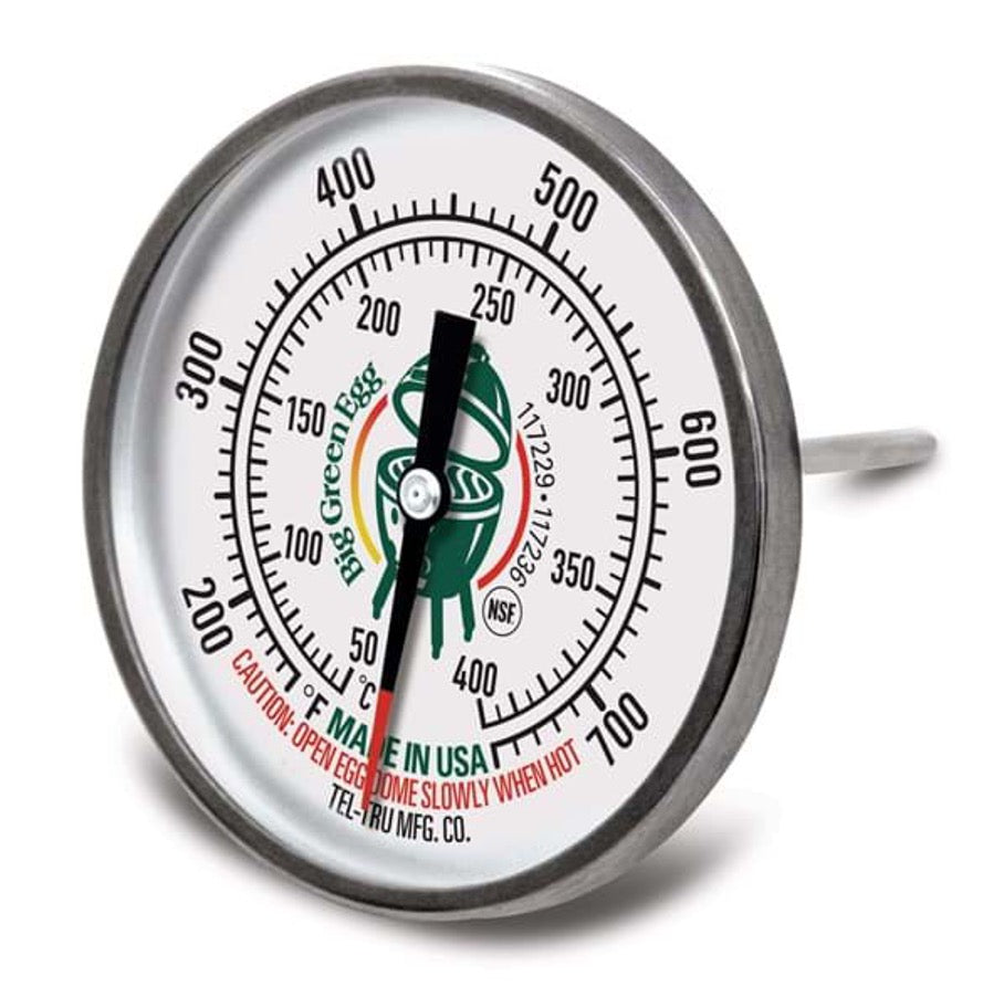 Een analoge temperatuurmeter van Big Green Egg. Het ronde display heeft een witte achtergrond met zwarte en rode markeringen die temperaturen aangeven van 50°F tot 750°F (of ongeveer 20°C tot 400°C). In het midden staat het Big Green Egg-logo afgebeeld. De thermometer heeft een metalen behuizing en een meetpen die in de barbecue wordt geplaatst om de interne temperatuur te meten. Onderaan staat de tekst "Made in USA" en een waarschuwing: "Open Egg dome slowly when hot". 