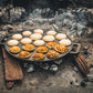 De gietijzeren pan op hetzelfde open vuur. Ditmaal met een volle pan, waarbij de bovenste helft van de poffertjes nog niet is omgedraaid en de onderste helft laat mooi goudbruin gebakken poffertjes zien.