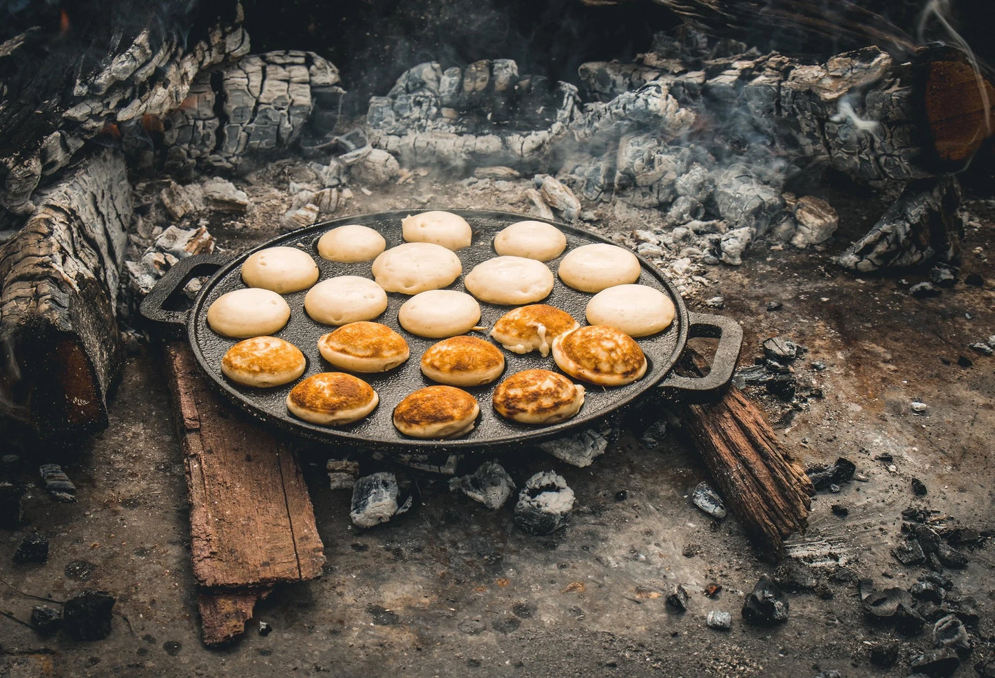 De gietijzeren pan op hetzelfde open vuur. Ditmaal met een volle pan, waarbij de bovenste helft van de poffertjes nog niet is omgedraaid en de onderste helft laat mooi goudbruin gebakken poffertjes zien.