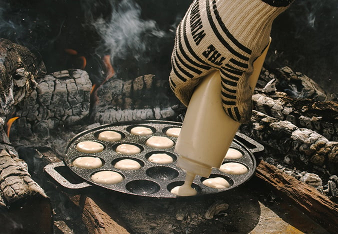 De gietijzeren poffertjespan naast een open vuur. Een hand spuit met een knijpfles het poffertjesbeslag in de pan.