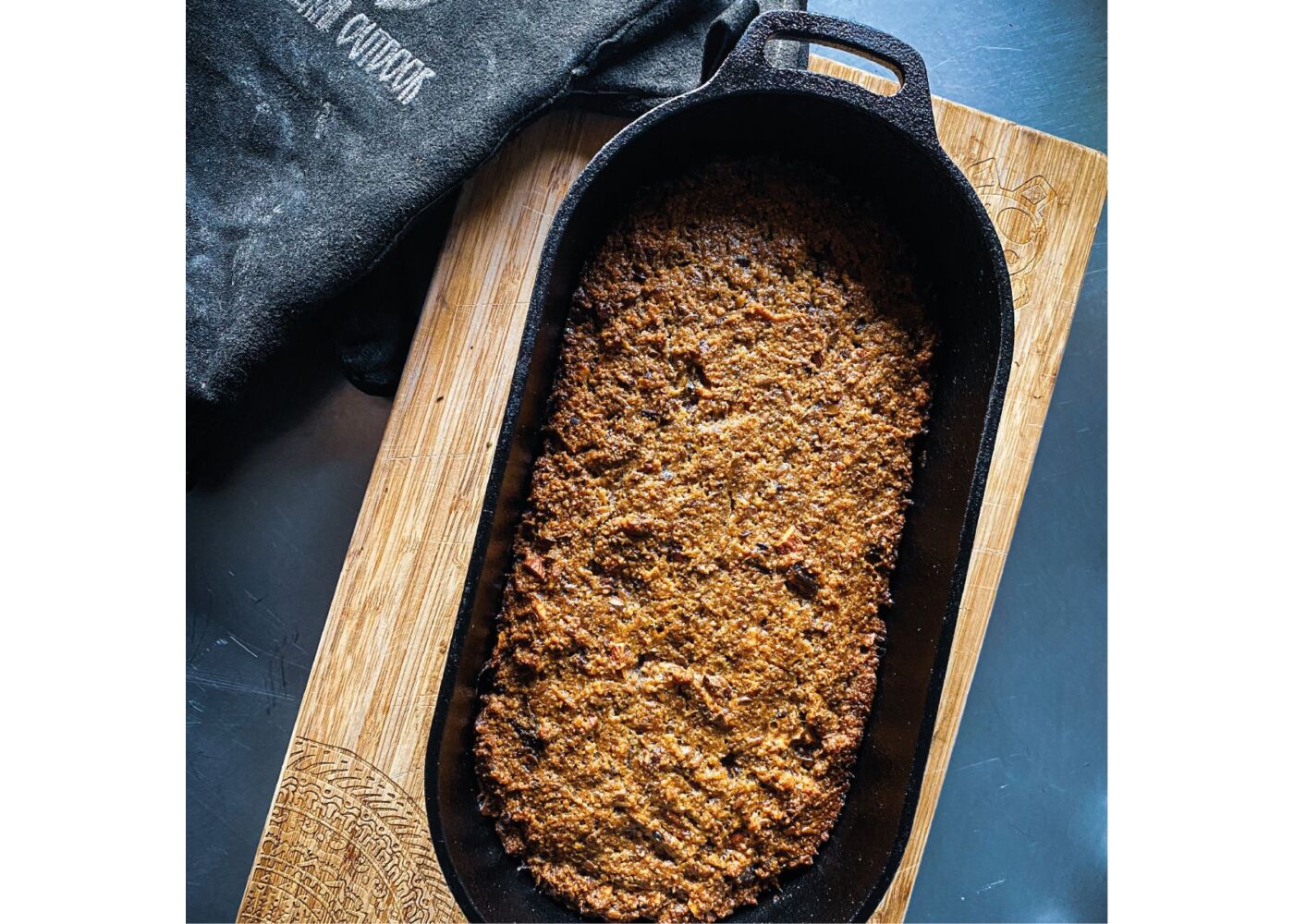 Een sfeerimpressie van de gietijzeren dutch oven zonder deksel. De dutch oven staat op een stoere houten snijplank op een donkerblauwe achtergrond. Naast de pan ligt een zwarte kookdoek.