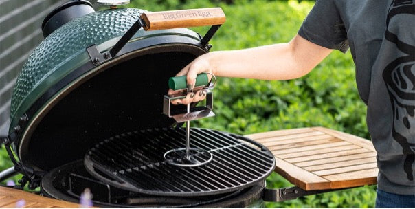 Iemand staat buiten bij een geopende Big Green Egg kamado en tilt met behulp van de grid lifter met één hand het gietijzeren rooster eruit. 