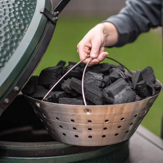 De met houtskool gevulde RVS mand staat op de rand van een Big Green Egg en wordt vastgehouden aan de handvaten door een mannenhand.