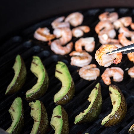 Close-up van het gietijzeren rooster in een Big Green Egg met daarop gegrilde garnalen en partjes avocado.